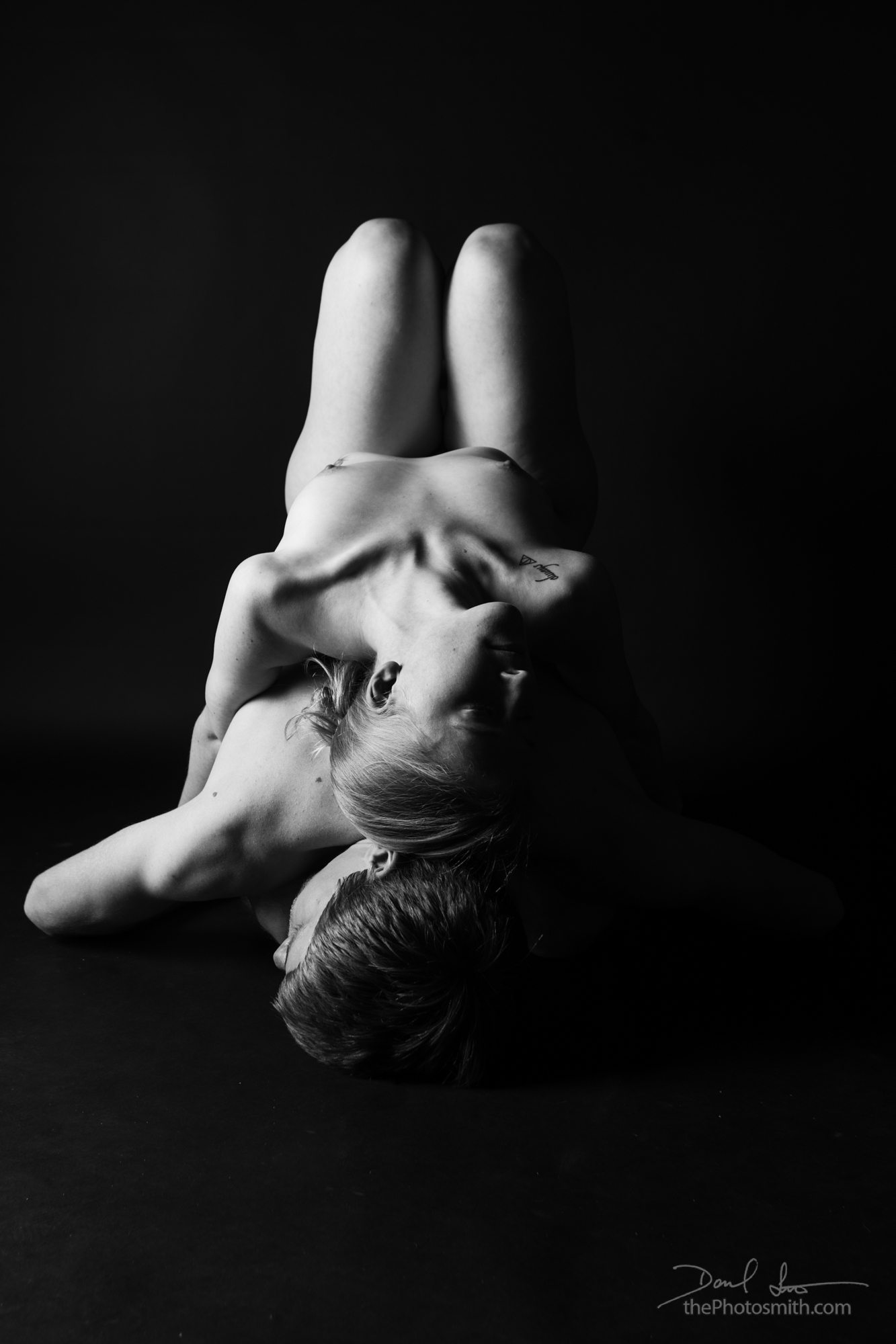 Black and white image of a nude woman laying atop a nude man, back to back.